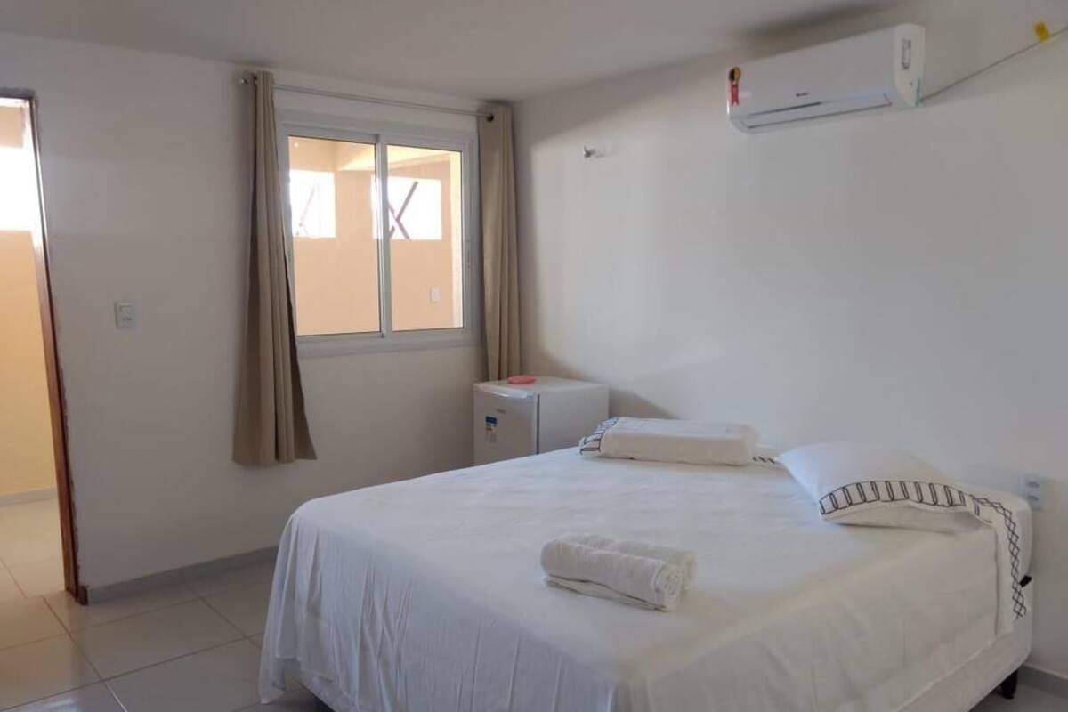 Una cama bien hecha con sábanas y almohadas blancas, una ventana con cortinas y un aire acondicionado instalado en la pared.