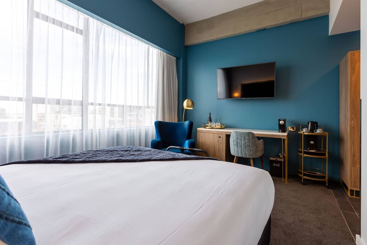 Habitación de hotel con una cama grande, un escritorio con silla, un televisor montado en la pared y una ventana con cortinas translúcidas.