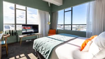 Une chambre d’hôtel avec un grand lit, un bureau avec une télévision, une chaise et une vue sur la ville grâce à de grandes fenêtres.