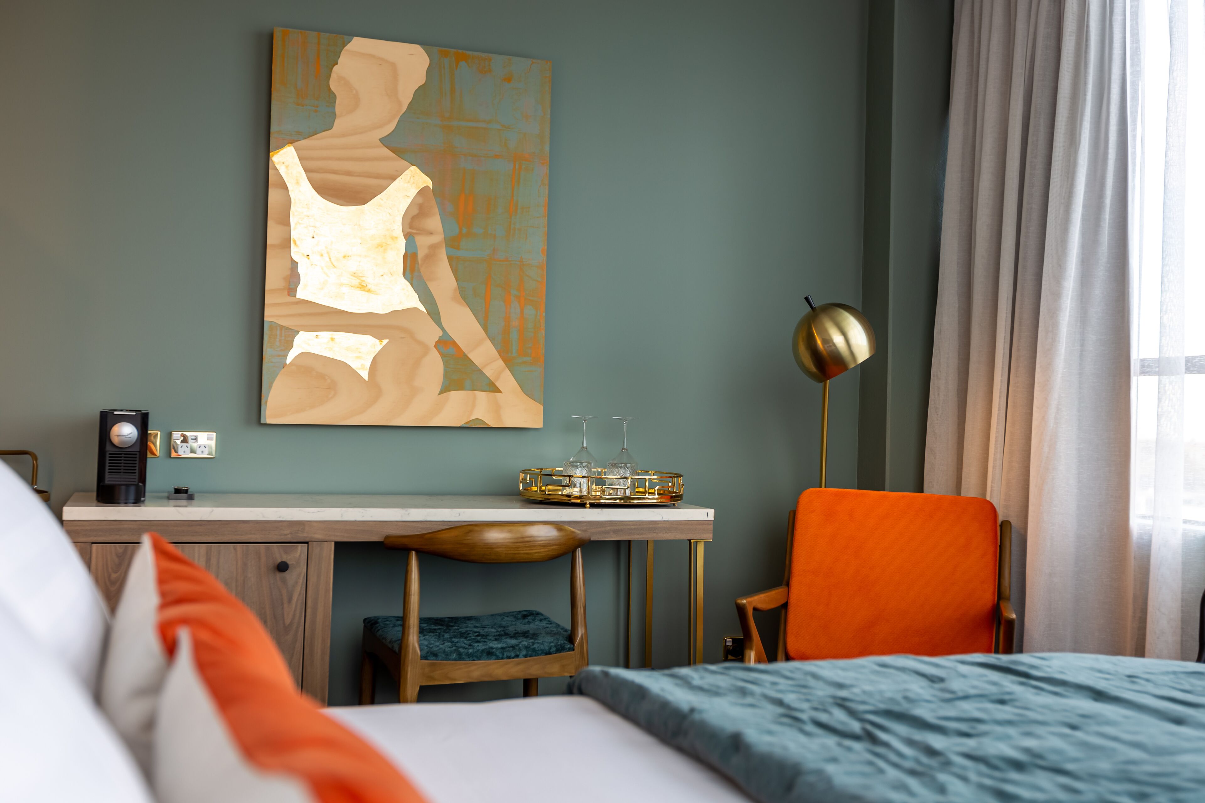 A hotel room with a bed, a desk, a chair, a lamp, and a painting on the wall.