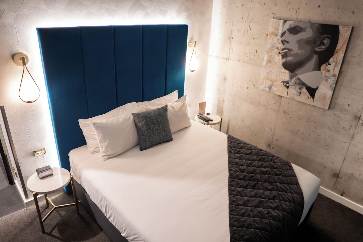 Una cama bien tendida con cabecera azul, una mesita de noche con lámpara y un cuadro en la pared.