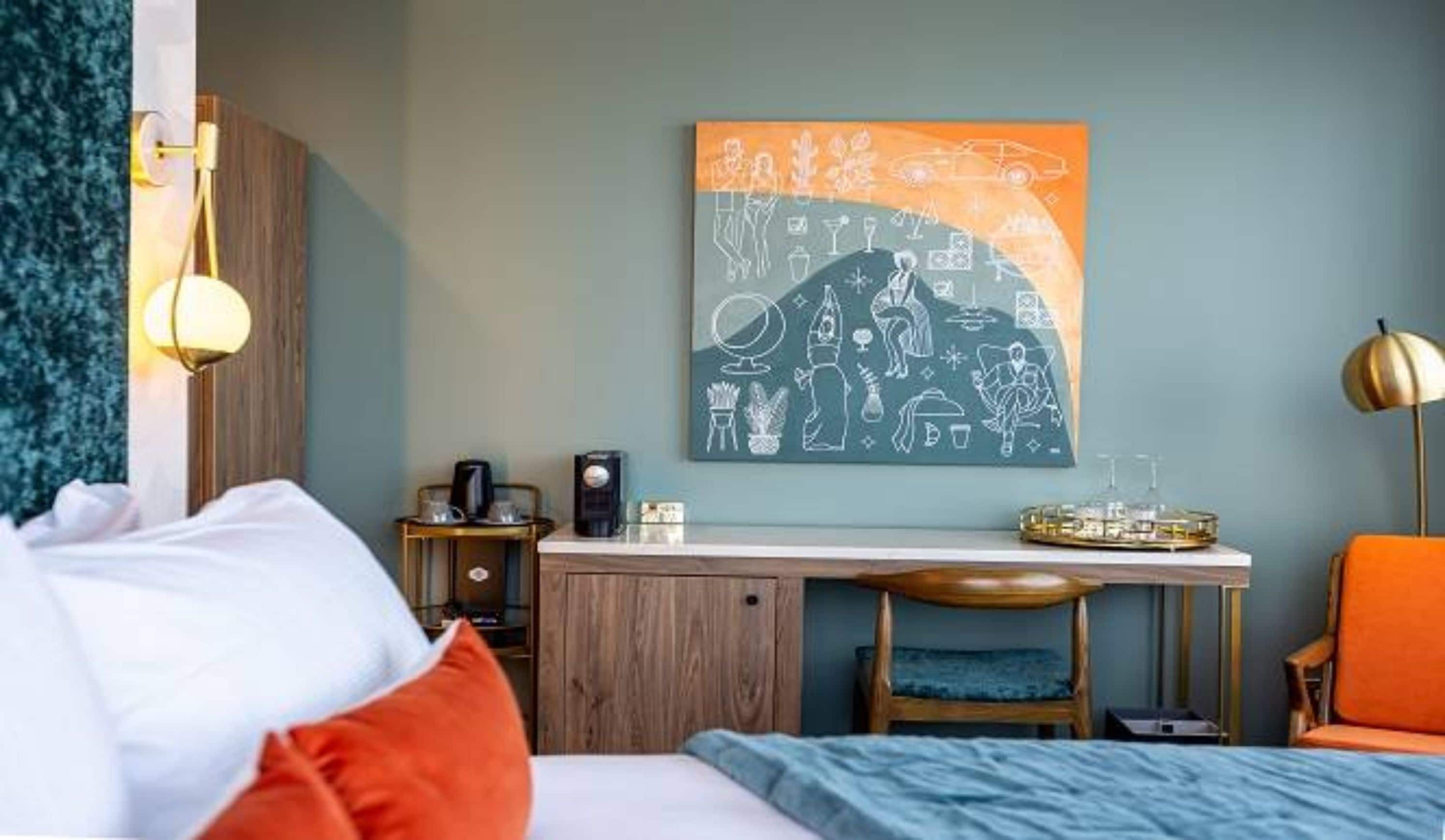 Habitación de hotel con cama, escritorio, silla, cafetera y una obra de arte en la pared.