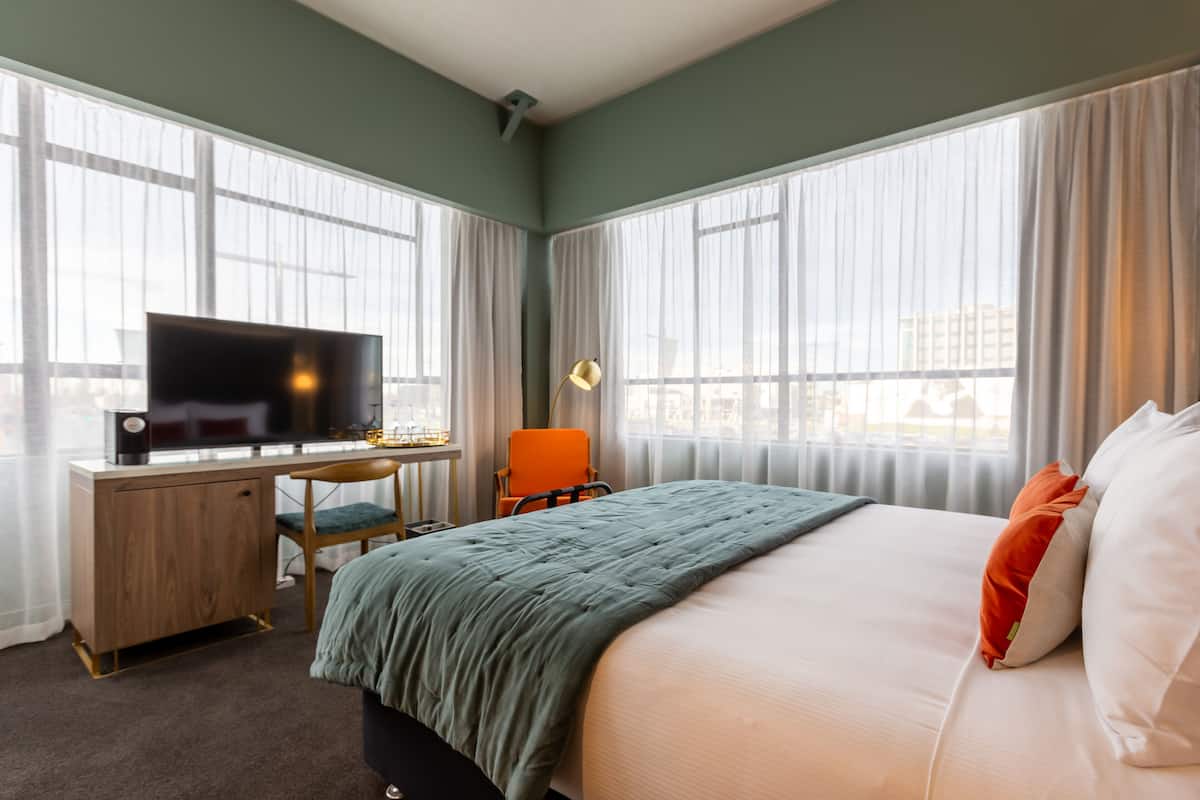 Habitación de hotel con cama, escritorio con televisor, una silla y ventanas amplias.