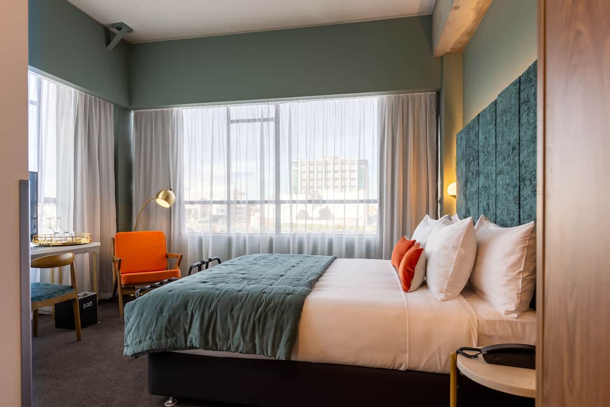Habitación de hotel con cama, escritorio, una silla naranja, lámpara y vistas a edificios a través de amplias ventanas.