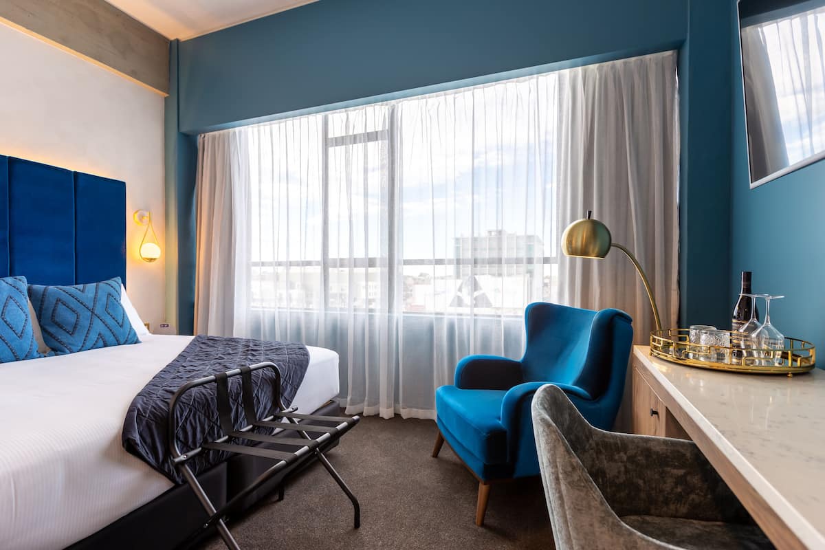 Una habitación de hotel con cama, escritorio, silla, lámpara y ventana con cortinas.