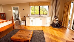 Amplia habitación con cama de madera, una mesita pequeña, zona de cocina con estufa y un amplio ventanal con vistas a zonas verdes.