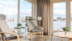 A hotel room with two grey lounge chairs, a small table with a lamp, and a view of the water and boats outside. - Amazing ship in Aalsmeer with harbor view (Aalsmeer)