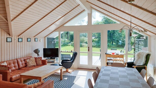 A spacious living room with a high ceiling, a television, a sofa, a dining table, and a view of a garden through large windows.
