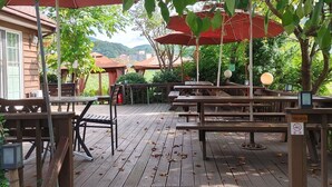 En træterrasse med picnicborde og parasoller, omgivet af træer og en bygning i baggrunden.