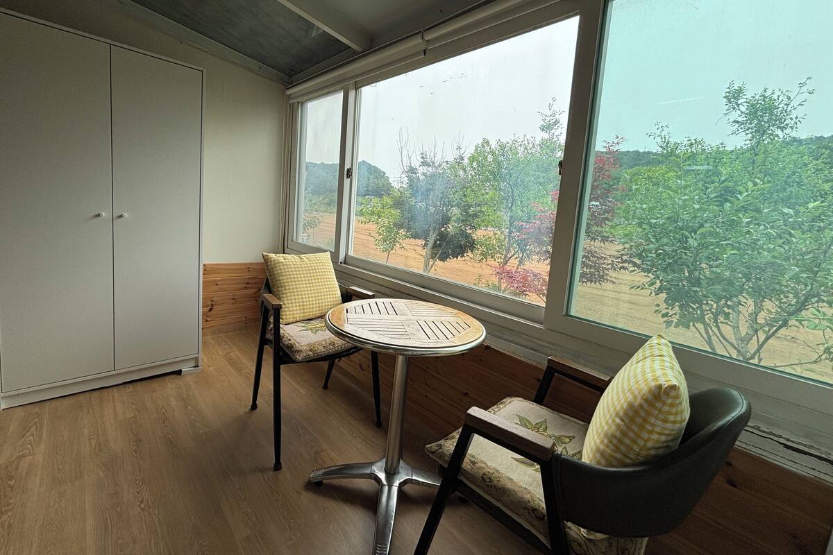Une pièce avec une grande fenêtre, une table ronde, deux chaises et une armoire.