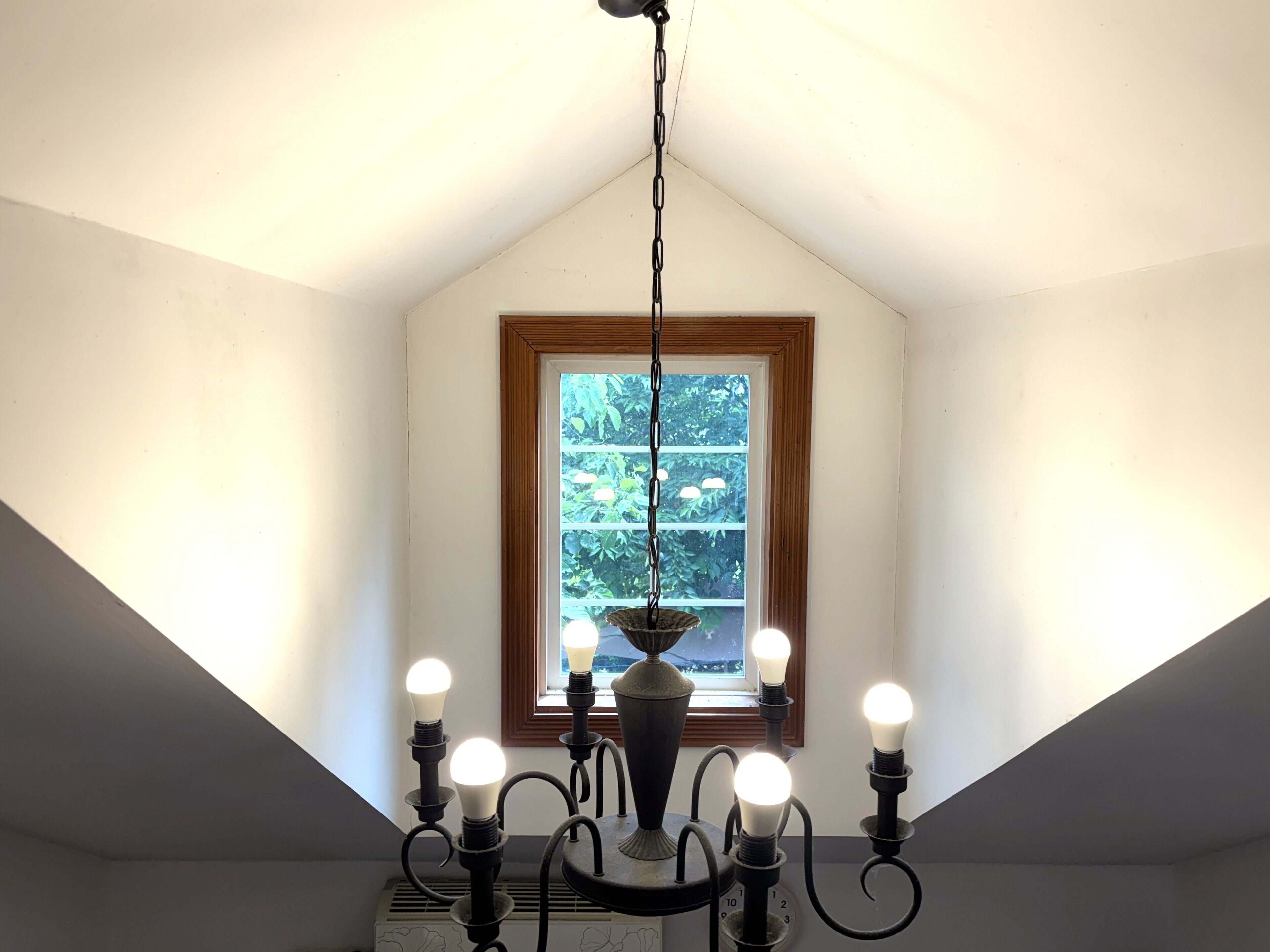 Una habitación con techo inclinado, ventana con vistas a la vegetación y un candelabro con varias bombillas.