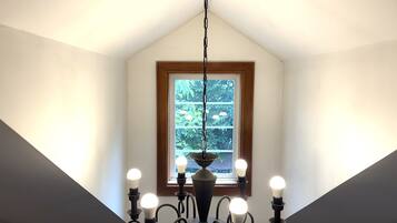 Una habitación con techo inclinado, ventana con vistas a la vegetación y un candelabro con varias bombillas.