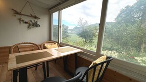 Spiseområde med et træbord og stole, store vinduer med udsigt til grøn bevoksning og træer.