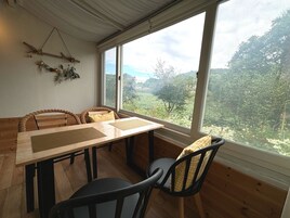 A dining area with a wooden table and chairs, large windows offering a view of greenery and trees.