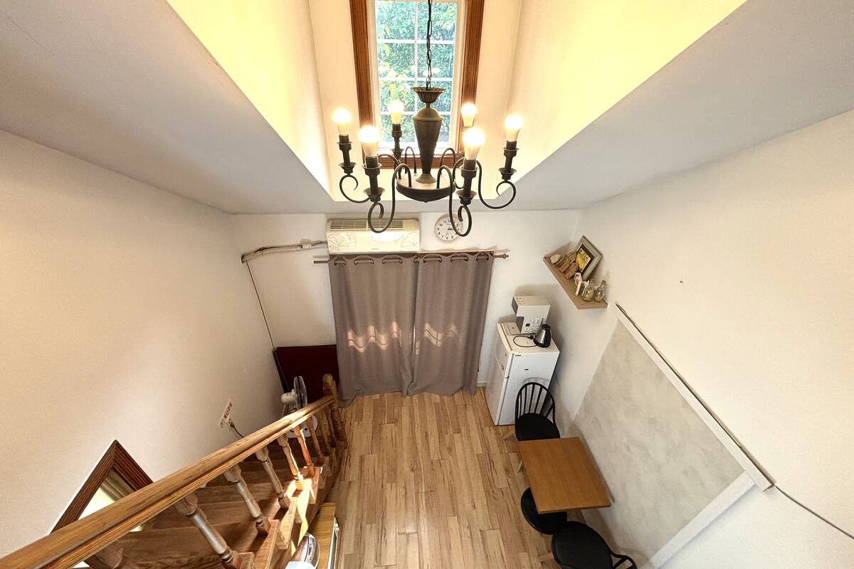 Un escalier menant à une chambre aménagée en mezzanine, dotée d’un plancher en bois, d’un lustre, d’un ventilateur de plafond, d’une fenêtre avec des rideaux et d’une petite table avec des chaises.