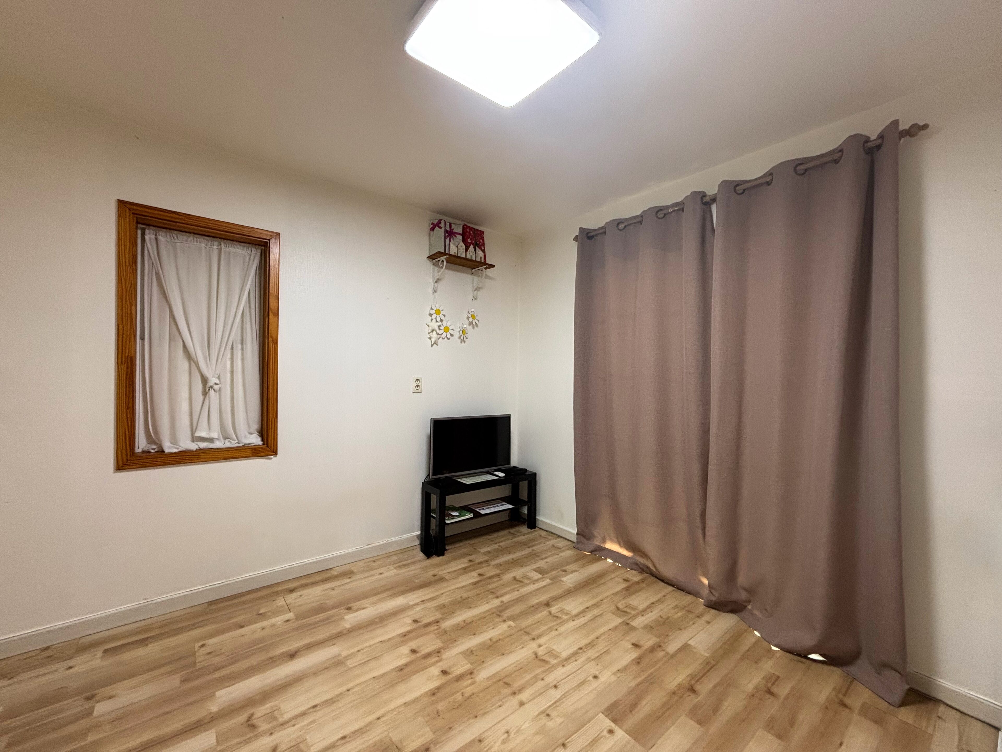 A room with wooden flooring, a television on a stand, a window with curtains, and a wall-mounted shelf with decorative items.