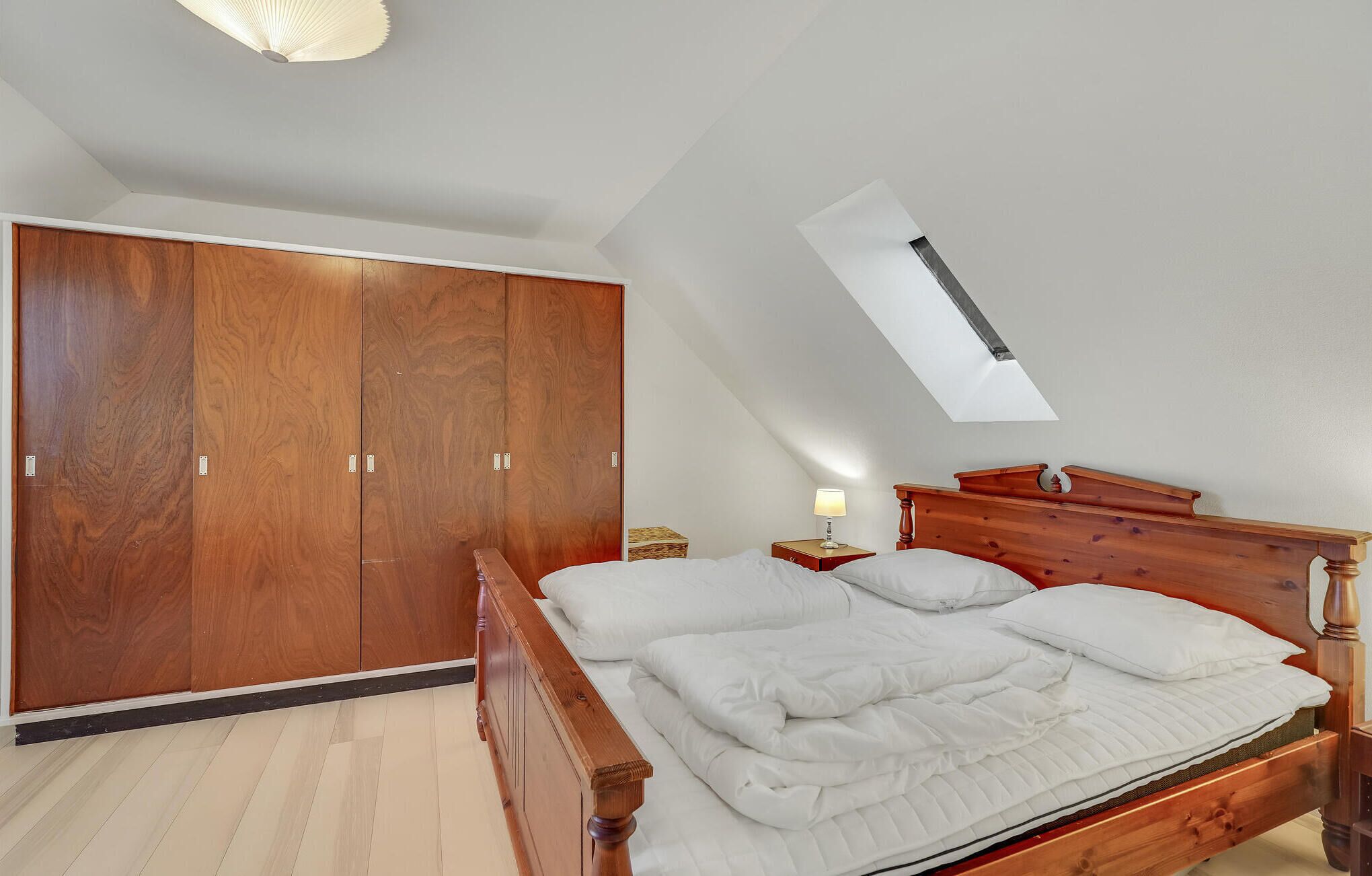 Un dormitorio con un armario de madera, una cama con ropa de cama blanca, una mesita de noche con una lámpara y un tragaluz.