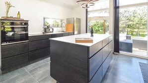 Une cuisine moderne dotée d’un grand îlot central, d’appareils électroménagers intégrés et offrant une vue sur l’extérieur.