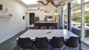 Une cuisine moderne dotée d’un îlot central, d’un coin repas avec une grande table et des chaises, et offrant une vue sur l’extérieur grâce à de grandes fenêtres.