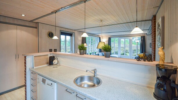 A modern kitchen with a stainless steel sink, wooden cabinets, a built-in dishwasher, and a window overlooking greenery. - Cozy home in Rømø with sauna (Rømø)