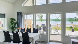 Zona pranzo con tavolo e sedie, ampie finestre con vista su un lago e un patio, e una pianta in vaso.