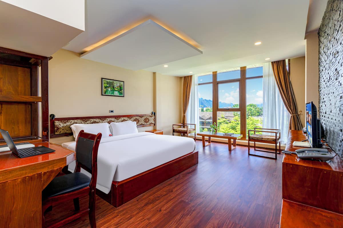 Ein Hotelzimmer mit einem großen Bett, einem Schreibtisch mit einem Laptop, einem Sessel, einem Fernseher und einem Fenster mit Blick ins Grüne.