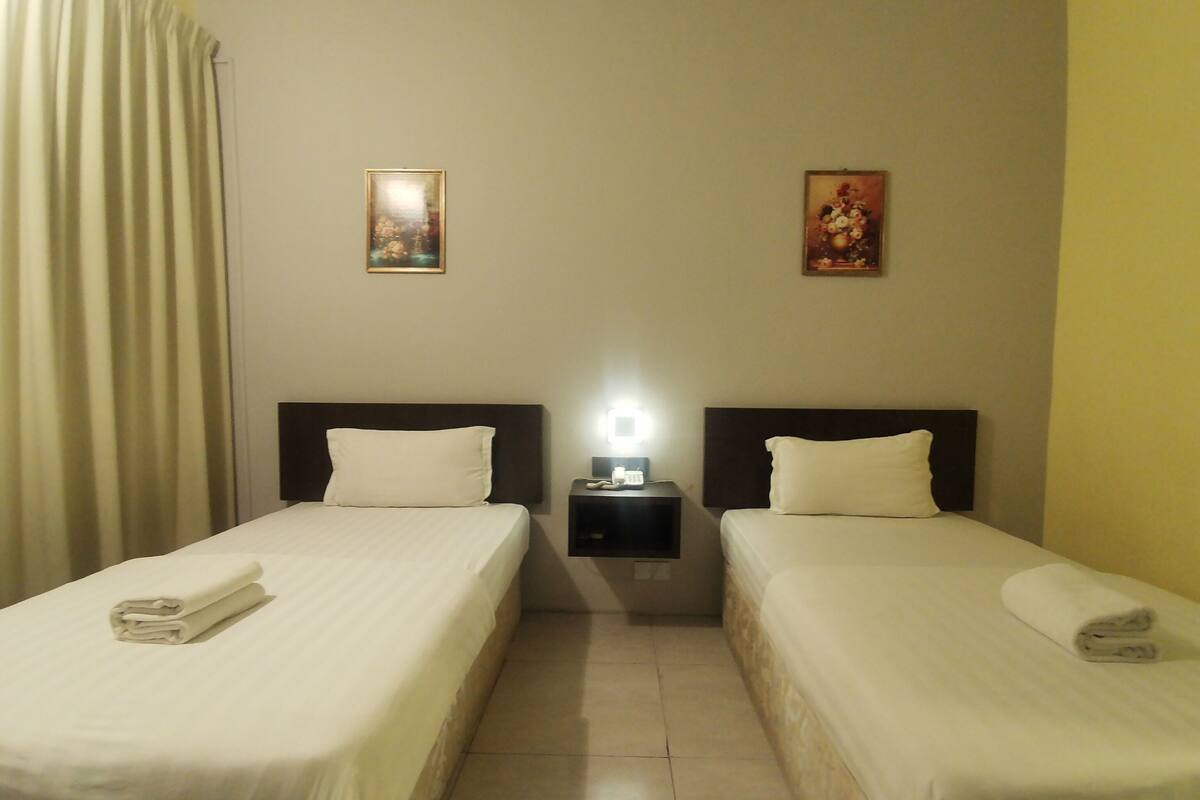 A hotel room with two single beds, a nightstand with a lamp, and two framed pictures on the wall.