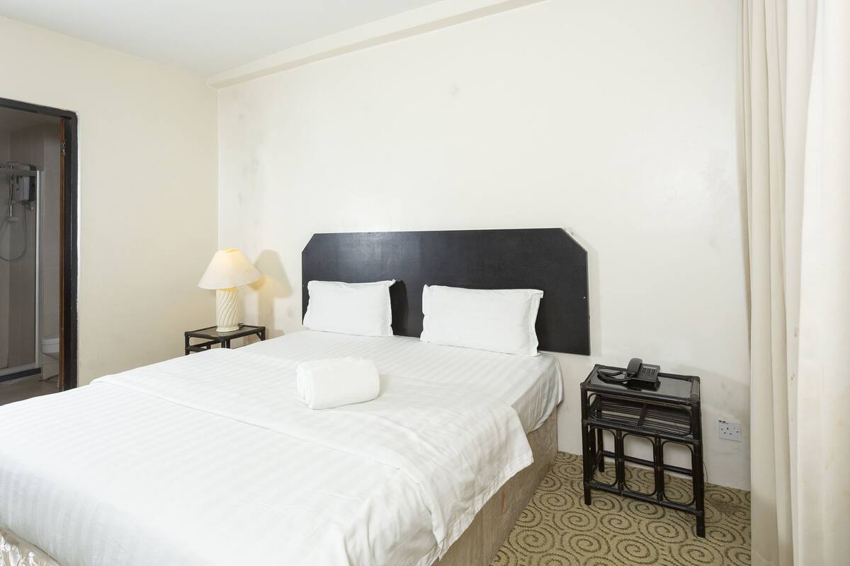 A hotel room with a bed, a nightstand with a lamp, a phone, and a shower area.