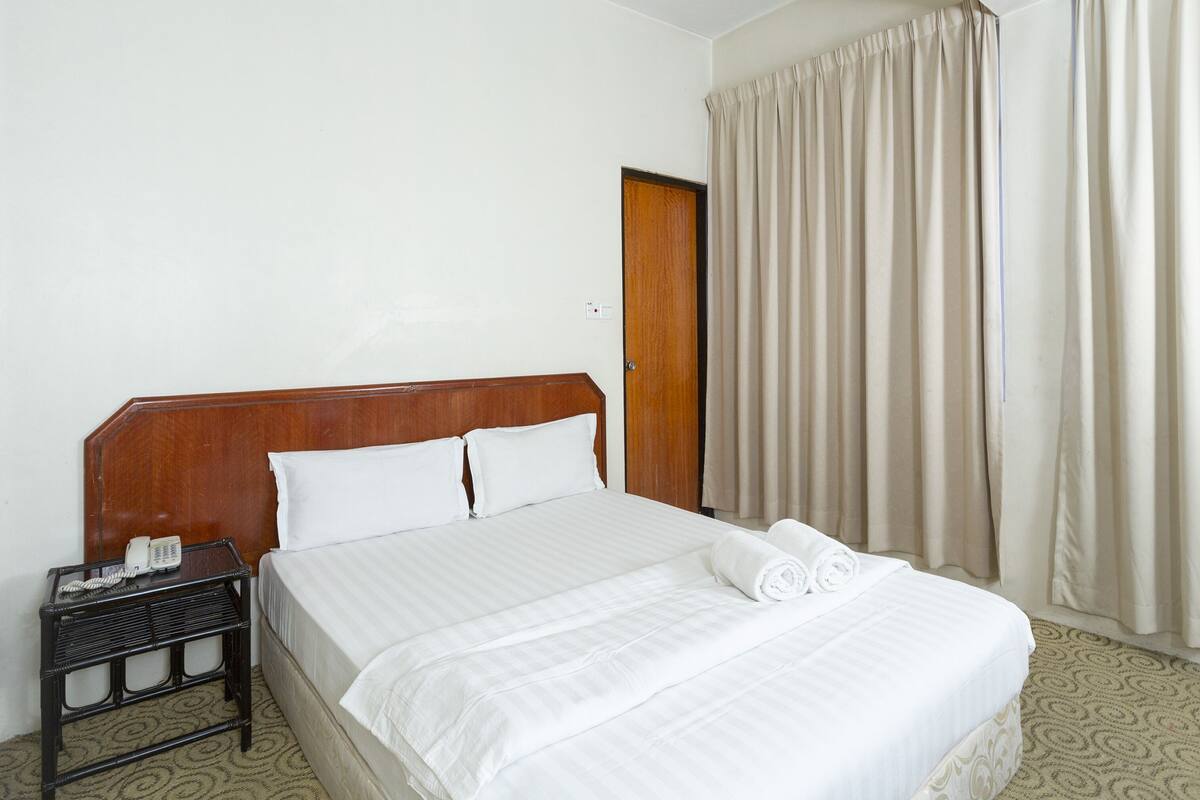 A hotel room with a wooden headboard, white bedding, a small bedside table with a telephone, and a window with curtains.