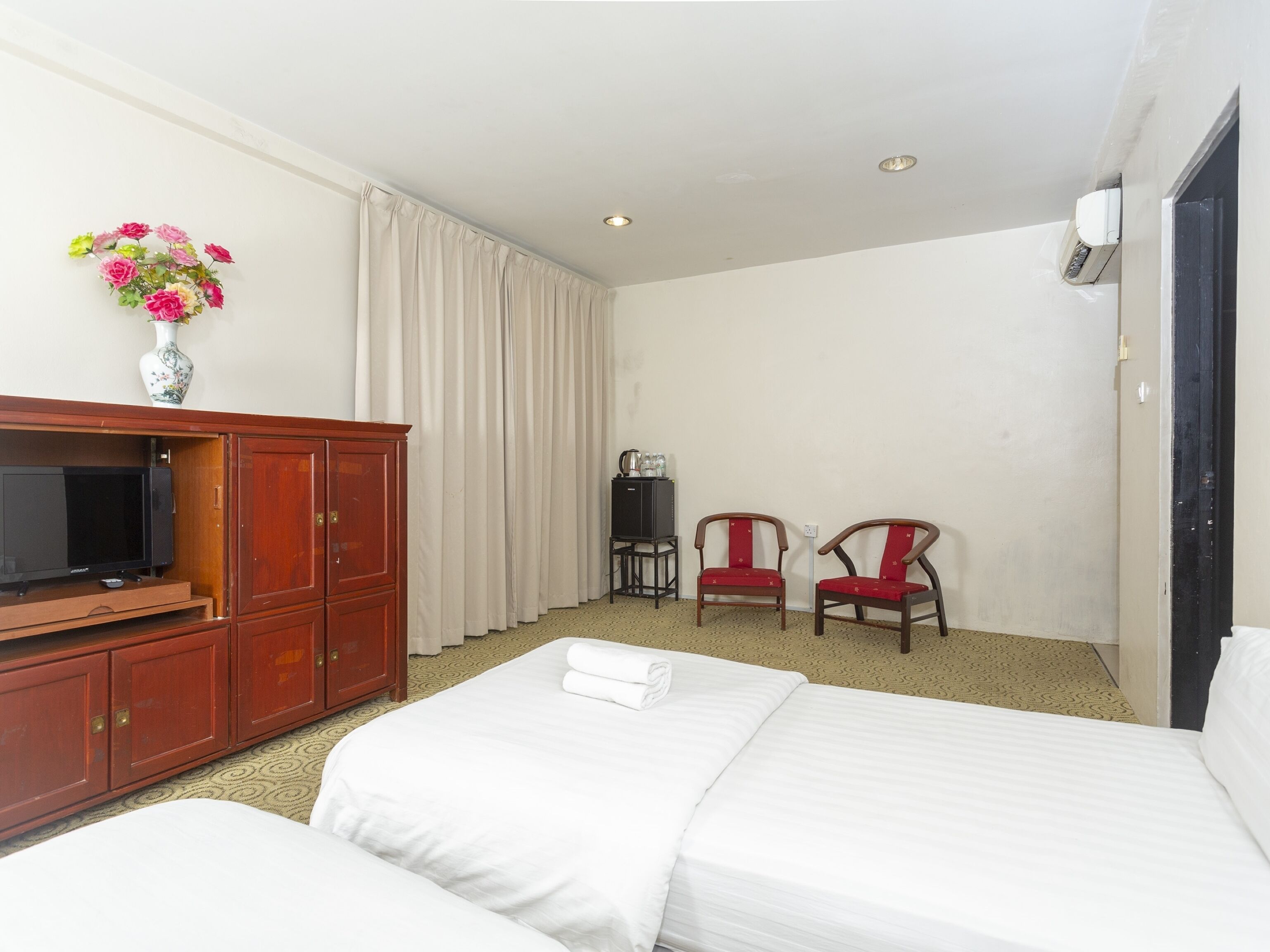 A hotel room with two beds, a TV on a wooden cabinet, a vase with flowers, two chairs, and a small table with bottles.