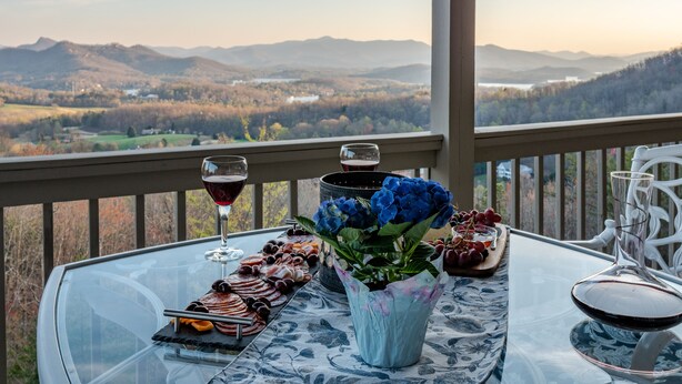 A table with a glass of red wine, a glass of white wine, a tray of assorted meats, a vase of blue flowers, and a bowl of grapes on a balcony with a scenic view of mountains and a lake.