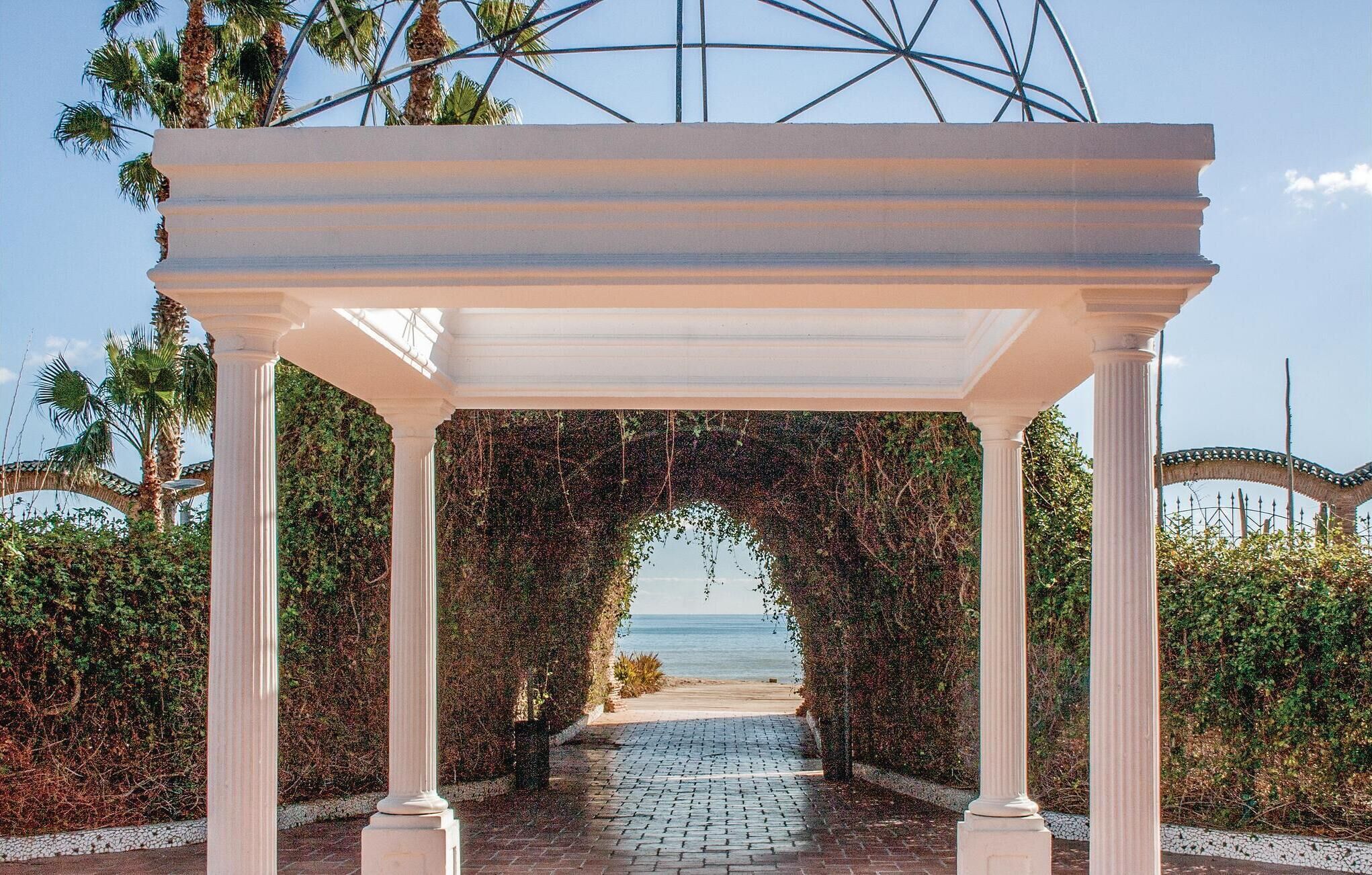 A covered walkway with white columns, leading to a view of the ocean.
