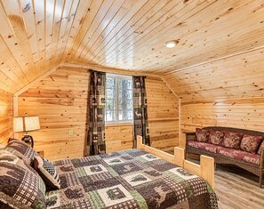 A cozy cabin room with a wooden ceiling, a bed with a patterned quilt, a window with curtains, and a wicker sofa.