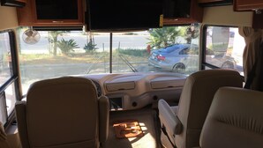 Een RV-interieur met twee grote schermen, een centrale gangpad en uitzicht op een geparkeerde auto.