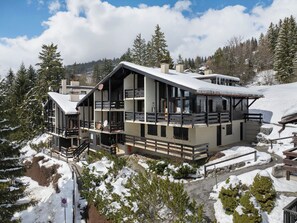 Un immeuble moderne à plusieurs étages, doté de balcons, entouré d’arbres couverts de neige et avec une chaîne de montagnes en arrière-plan.