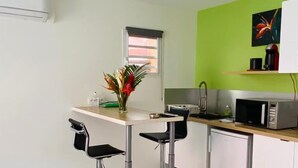 Cozinha moderna com armários brancos, uma parede verde de destaque, micro-ondas e um refrigerador pequeno. Há dois banquetas pretas e um vaso com flores na bancada.