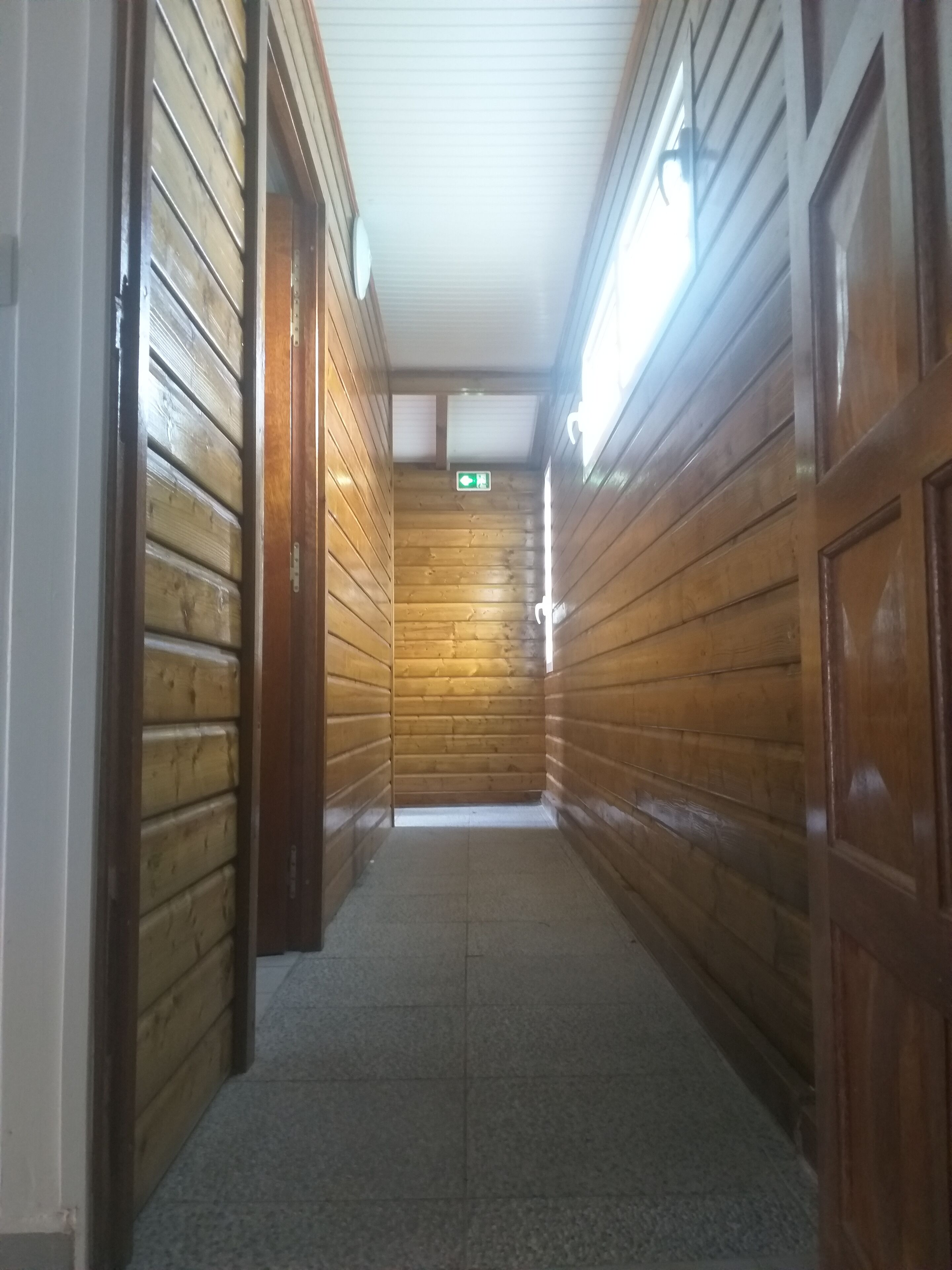 A narrow hallway with wooden walls and a tiled floor, leading to a door with a green emergency exit sign.
