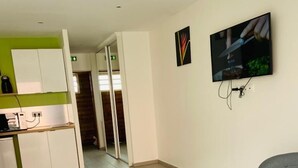 Quarto de hotel moderno com cama, poltrona cinza, televisão, cozinha compacta e porta deslizante para um banheiro.