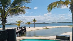 Zona de piscina con terraza de madera, palmeras y vista a una playa y al agua.