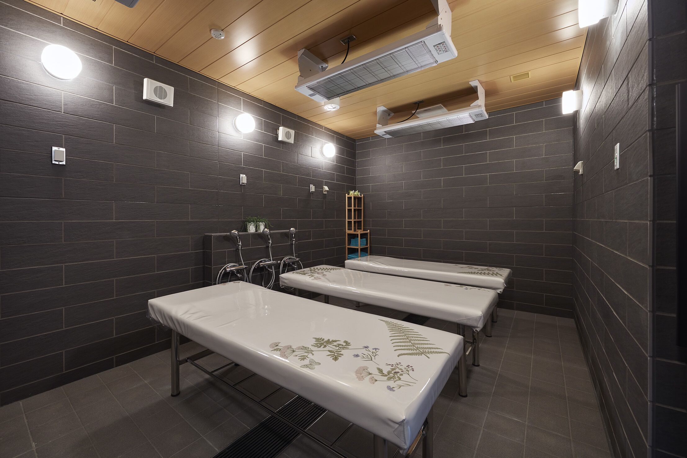 Una habitación con paredes de baldosas oscuras, tres camas blancas con diseños florales y unidades de aire acondicionado en el techo.