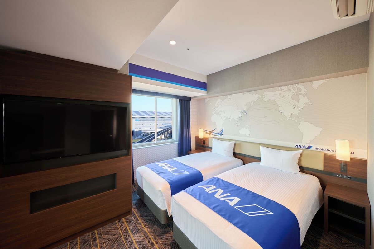 Ein Hotelzimmer mit zwei Betten, einem Fernseher, einer Weltkarte an der Wand und einem Fenster mit Blick auf ein Gebäude.