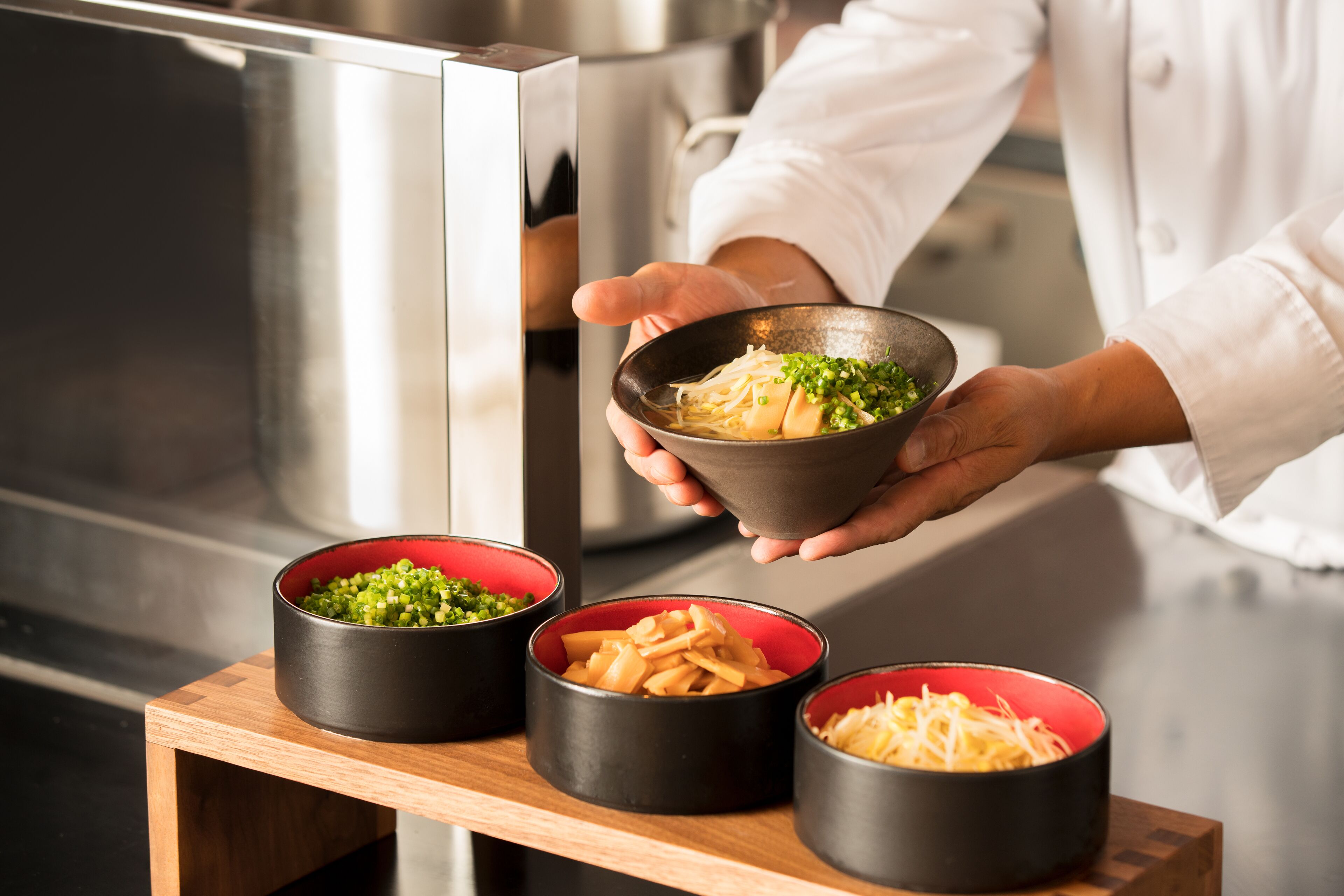 A chef holding a bowl of noodles with green toppings, with three other bowls of different ingredients on a wooden stand.