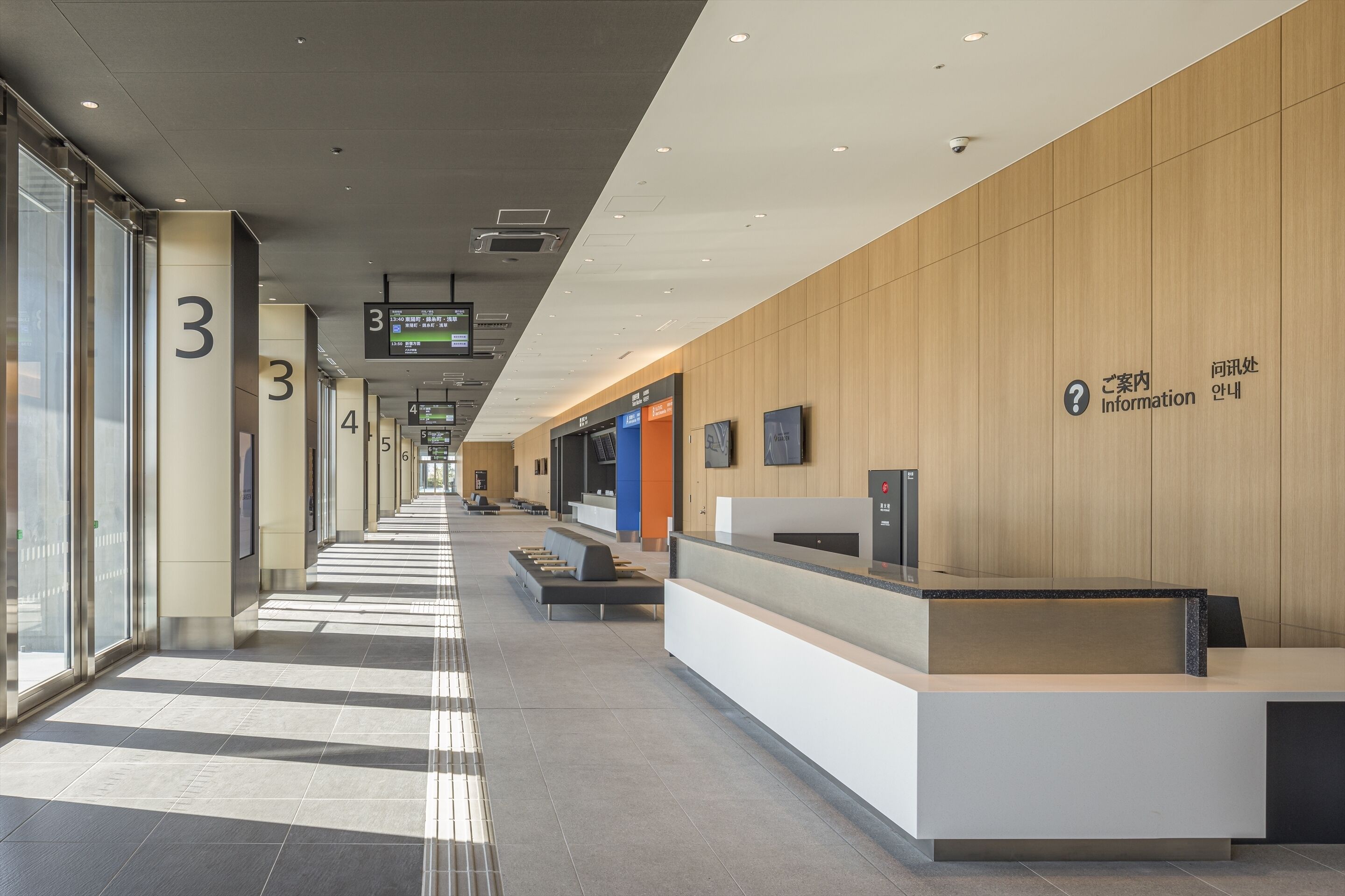 A modern hotel reception area with a long corridor, numbered pillars, and a reception desk with a digital display.