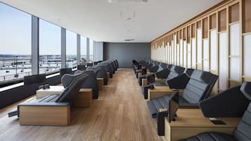 A modern hotel room with a long row of black leather sofas and wooden tables, large windows offering a view of the airport, and a wooden partition wall.