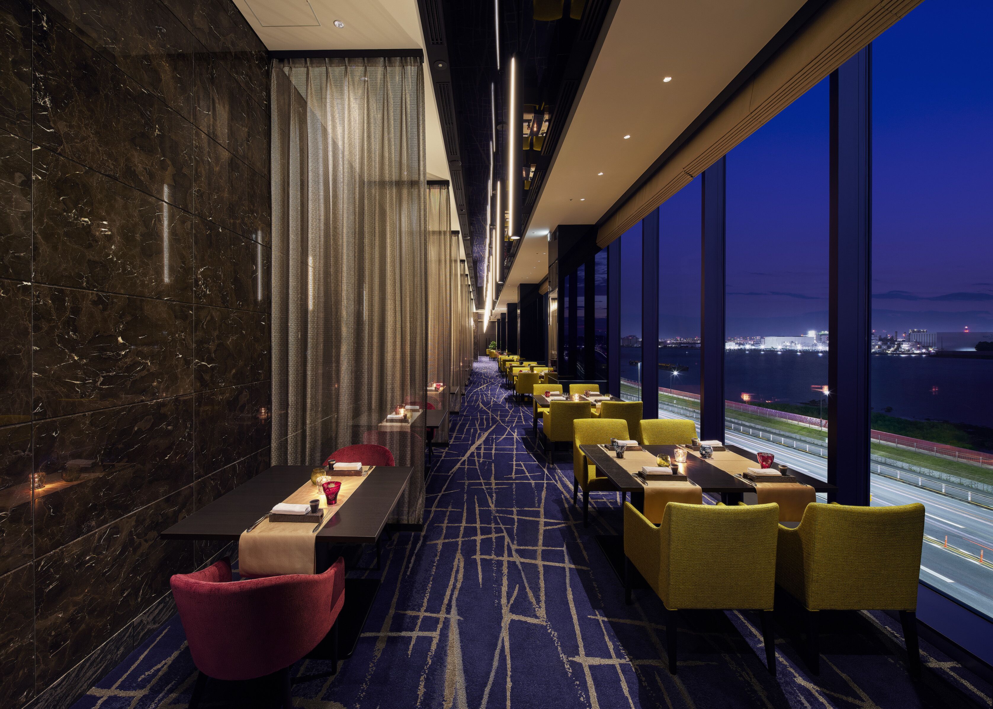 A modern hotel lobby with a long table and chairs, a patterned carpet, and large windows with a view of a cityscape and water.