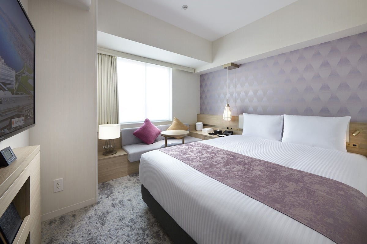 Ein Hotelzimmer mit einem großen Bett, einem Nachttisch, einem Fenster mit Vorhängen, einem an der Wand befestigten Fernseher und einem Teppichboden.
