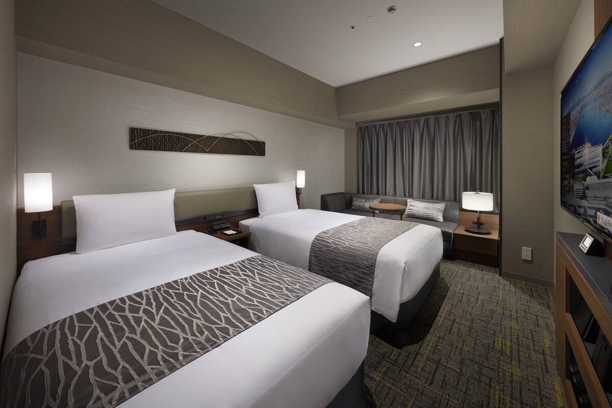 Ein Hotelzimmer mit zwei Betten, einem Schreibtisch, einem Fernseher und einem großen Fenster mit Vorhängen.