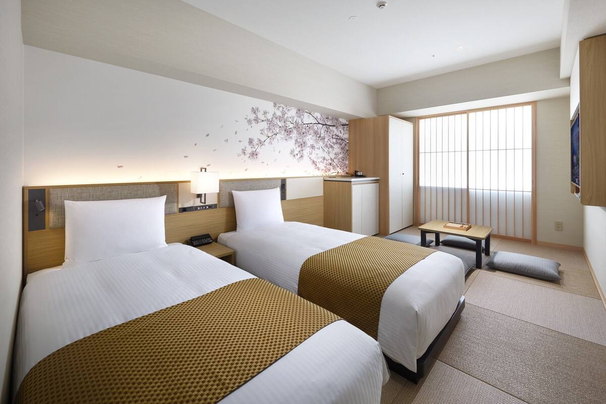 Ein Hotelzimmer mit zwei Betten, einem kleinen Schreibtisch, einem an der Wand befestigten Fernseher und einem großen Fenster mit Jalousien.