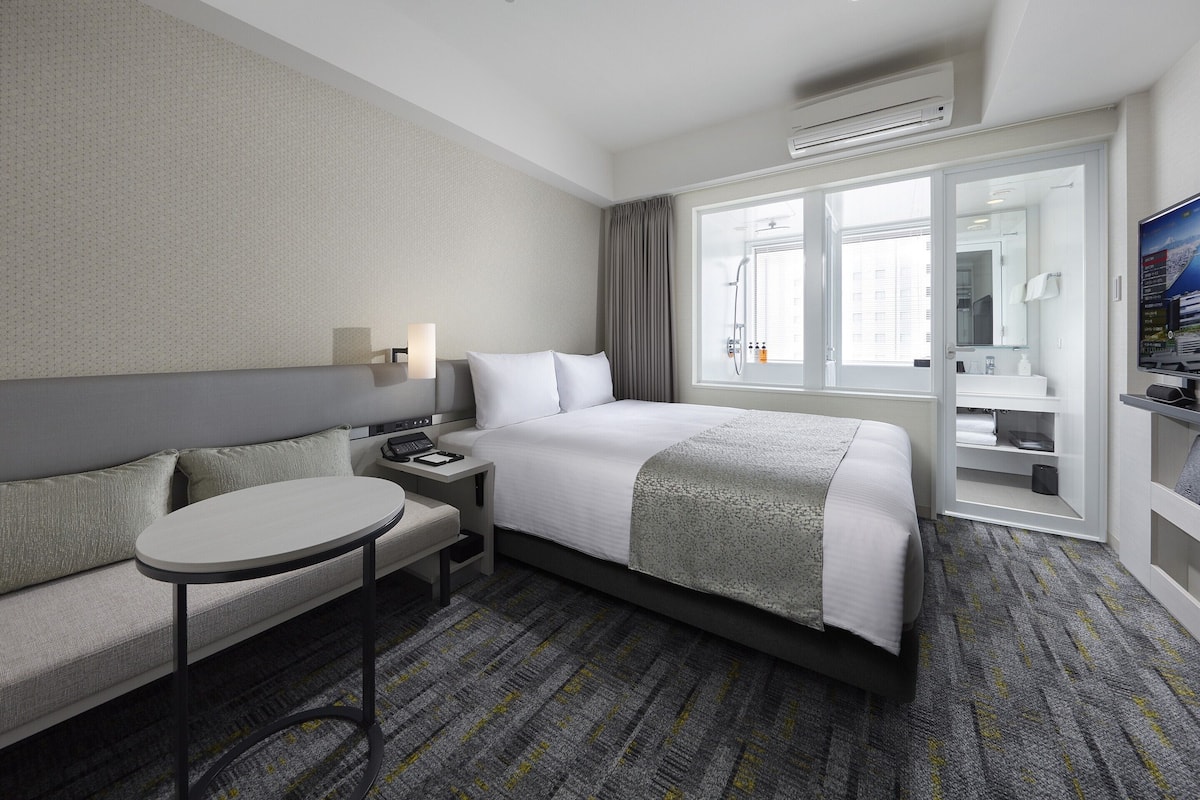 Ein Hotelzimmer mit Bett, einem kleinen Tisch, einer Couch, einer Dusche und einem Fernseher.