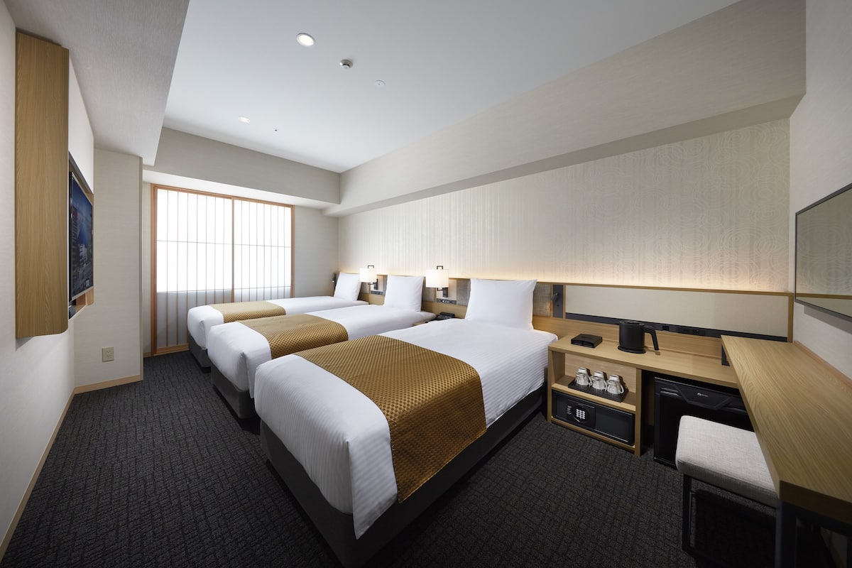 Ein Hotelzimmer mit zwei Betten, einem Schreibtisch, einem Sessel, einem kleinen Tisch, einem Nachttisch, einer Lampe, einer Kaffeemaschine und einem Fernseher.